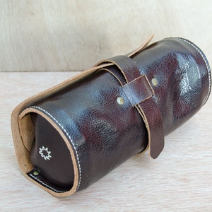 May include: A brown leather roll-up case with a tan leather strap and a white star design. The case is closed and lying on a light wood surface.