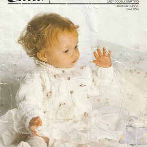 Może przedstawiać: Biały dzianinowy kardigan dla niemowlęcia z wzorem warkoczowym i różowymi kwiatowymi ozdobami. Kardigan zapinany jest z przodu na guziki i ma długie rękawy. Kardigan jest noszony przez niemowlę siedzące na białym kocu. Tekst "8775A BABY DOUBLE KNITTING 48-56 cm 19-22 in. Four sizes BONNY BABY 1988" jest wydrukowany na stronie.