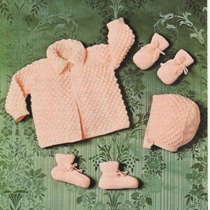 May include: A pink knitted baby cardigan, hat, and booties set. The cardigan has a button closure and a collar. The hat has a pointed top. The booties have a tie at the top. The set is laid out on a green floral background. The text "Baby Golden Sovereign Q.K. 1022 PRICE CODE A 17"-19" 43-48 cm" is printed on the top of the image.