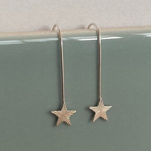 Puede incluir: Un par de pendientes de estrella plateados. Cada pendiente presenta un gancho largo y delgado y un dije en forma de estrella con detalles grabados. Fondo azul verdoso apagado.