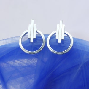 May include: A pair of silver earrings with a geometric design. Each earring features a circle with a vertical line pattern inside.