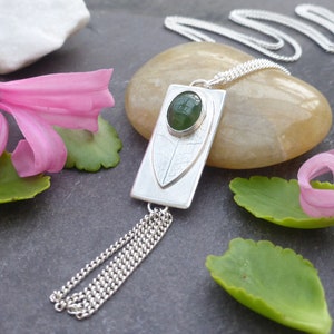 Puede incluir: Un collar con colgante de plata con un colgante rectangular con una piedra preciosa verde y un diseño de hoja. El collar tiene una cadena y detalles de borlas. Flores rosas y hojas verdes en el fondo.
