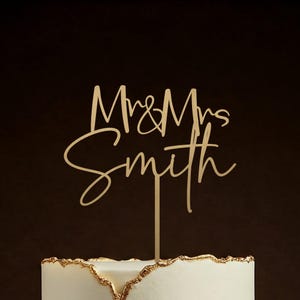 May include: A wooden cake topper reading "Mrs & Mrs Smith" in a cursive script. The topper is set on a white cake with gold edging. The backdrop is a dark brown colour.