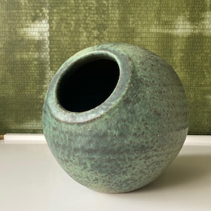 May include: A spherical ceramic vase in green and brown hues. The vase features a wide opening and a textured surface. The interior is dark, contrasting with the speckled exterior. A distinctive decorative item, suitable for display or as a functional piece.