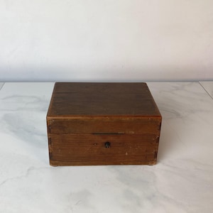 Puede incluir: Una caja de madera con tapa abatible y un pequeño pestillo de metal. La caja es de madera oscura y tiene un acabado natural.