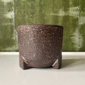 Può includere: Un vaso in ceramica marrone scuro e strutturato, di forma cilindrica. Il vaso ha una superficie ruvida e macchiettata e poggia su tre piccoli piedini triangolari. Lo sfondo presenta una parete verde strutturata.