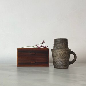 Op de afbeelding: Een houten doos met twee laden met een donkere houtkleur en een keramische vaas met een gespikkeld grijs glazuur.