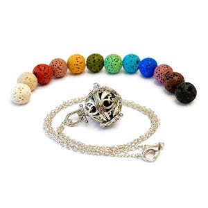 May include: A silver-toned metal locket pendant on a delicate chain, accompanied by a set of twelve porous, spherical lava beads in various colors including white, red, yellow, green, blue, brown, and black, arranged in a curved line.