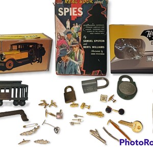 May include: A collection of vintage items including a small toy car, a toy train, a book titled "Real Book About Spies", a small box with a yellow lid, a small white bust, a key, a set of cufflinks, a set of lock and keys, and a small box with the text "Hoodies Collectible Auto Ornaments Lootcrate Exclusive".