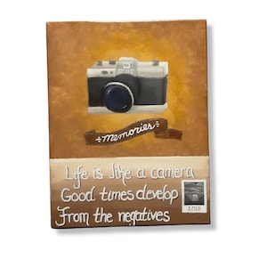 May include: A painting of a vintage camera with the text "SMILE" on it. The camera is on a brown background with a ribbon that says "Memories." Below the camera is the text "Life is like a camera, Good times develop from the negatives." There is a small photo below the text with the year 2018.