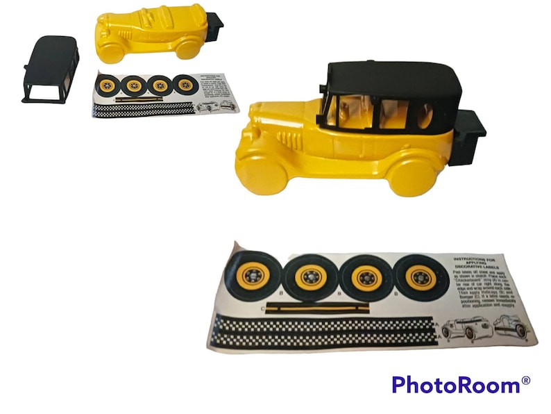 May include: A yellow toy car with a black roof and a black boot. The car has four wheels and is missing one of its wheels. The car is on a white background. The car is a vintage style car.