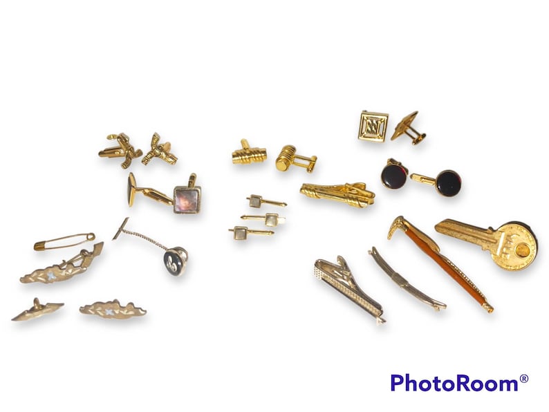 May include: A collection of gold-toned cufflinks, tie clips, and a key-shaped pin. The cufflinks feature various designs, including a square with an initial, a pair with a mother-of-pearl inlay, and a pair with a geometric design. The tie clips are in various shapes, including a straight bar, a curved bar, and a key shape. The key-shaped pin has a detailed design and a gold-toned finish.