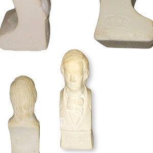 May include: White plaster bust of a man with short hair and a bow tie. The bust is on a base with the word "SCHUMANN" inscribed on it.
