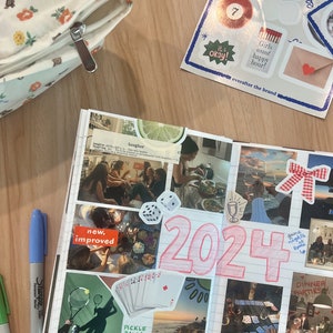 May include: A pink and white journal with a collage of photos and stickers. The journal has the number 2024 written in pink on the front. The photos include a group of people laughing, a game of cards, and a sunset over the ocean. The stickers include a red heart, a red bow, and a glass of wine.