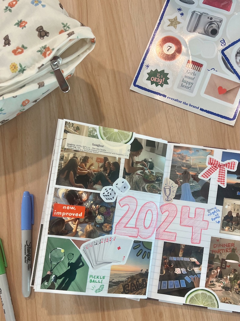 May include: A pink and white journal with a collage of photos and stickers. The journal has the number 2024 written in pink on the front. The photos include a group of people laughing, a game of cards, and a sunset over the ocean. The stickers include a red heart, a red bow, and a glass of wine.