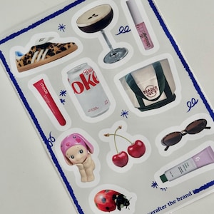 May include: A sheet of stickers featuring various designs, including a leopard print sneaker, a cocktail glass, a can of Diet Coke, a Trader Joe's tote bag, a cherry, a ladybug, a doll, sunglasses, a tube of lip gloss, and a tube of hand cream. The sheet is bordered in blue and white stars.