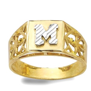 May include: A gold ring with a square-shaped setting featuring the letter "M" in white metal. The ring has a delicate, intricate design around the setting.