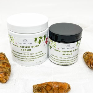 Puede incluir: Dos tarros de exfoliante corporal con tapas blancas y etiquetas negras. Las etiquetas tienen un diseño floral y el texto "NATUREL BLISS EMULSIFIED BODY SCRUB TURMERIC BENTONITE CLAY/BAOBAB". Los tarros están sobre una superficie blanca con dos trozos de raíz de cúrcuma.