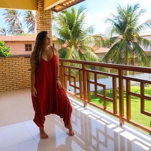 May include: A woman wearing a red jumpsuit stands on a balcony overlooking a tropical landscape. The balcony has a wooden railing with a geometric design. Palm trees and lush greenery are visible in the background.