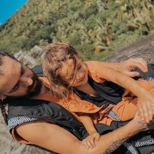 May include: A person wearing a black vest with a geometric pattern and an orange shirt, holding a child wearing an orange outfit. The background features a lush green hillside and a blue sky.