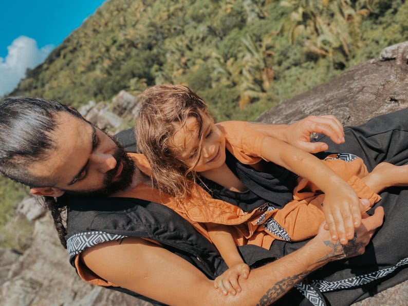 May include: A person wearing a black vest with a geometric pattern and an orange shirt, holding a child wearing an orange outfit. The background features a lush green hillside and a blue sky.