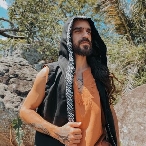May include: A black hooded vest with a white and black geometric patterned trim. The vest is worn over an orange tunic with a fringed hem. The person is outdoors, surrounded by trees and rocks.