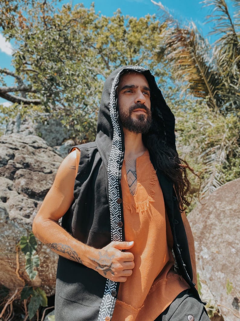 May include: A black hooded vest with a white and black geometric patterned trim. The vest is worn over an orange tunic with a fringed hem. The person is outdoors, surrounded by trees and rocks.
