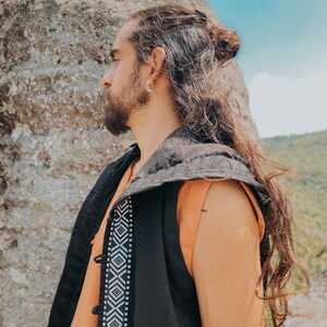 May include: A black vest with a geometric white pattern down the front, worn over an orange top. The vest has a hood and button closures. The person has long, wavy brown hair and is wearing a silver earring.