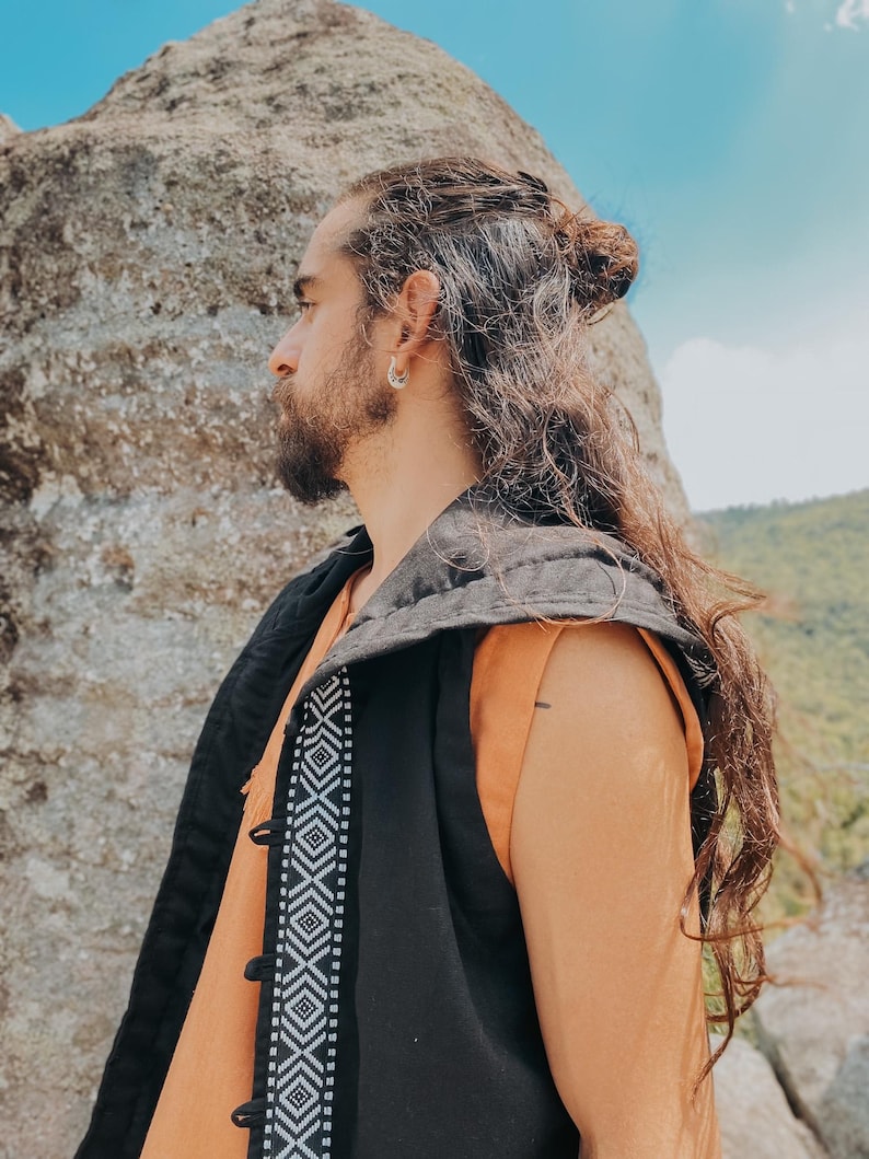 May include: A black vest with a geometric white pattern down the front, worn over an orange top. The vest has a hood and button closures. The person has long, wavy brown hair and is wearing a silver earring.