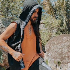 May include: Black hooded vest with white geometric trim, worn over an orange top. The vest has a button closure. The outfit is paired with black pants with matching geometric trim. The clothing has a bohemian style.