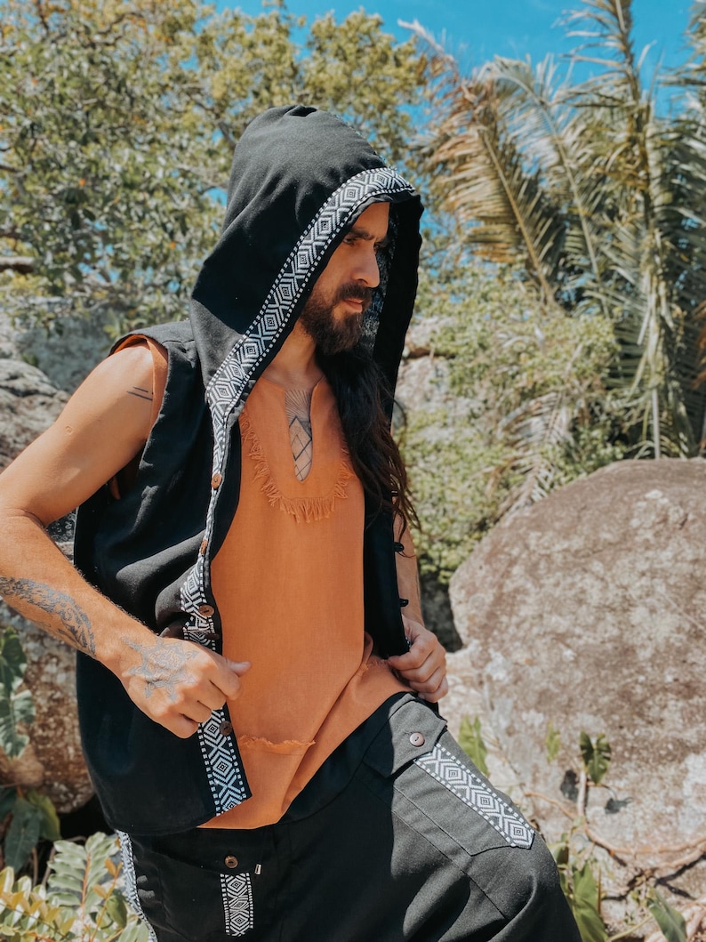 May include: Black hooded vest with white geometric trim, worn over an orange top. The vest has a button closure. The outfit is paired with black pants with matching geometric trim. The clothing has a bohemian style.