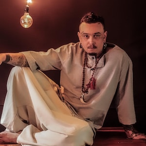 May include: A person wearing a light gray linen shirt and off-white pants, seated on a wooden surface. The person is adorned with jewelry and tattoos. A light bulb hangs from above, casting a warm glow.