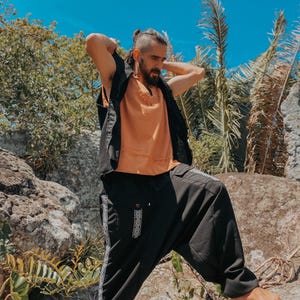 May include: A person wearing an orange top, black vest, and black pants with white patterned accents. The person is standing on rocks outdoors with a blue sky and greenery in the background.