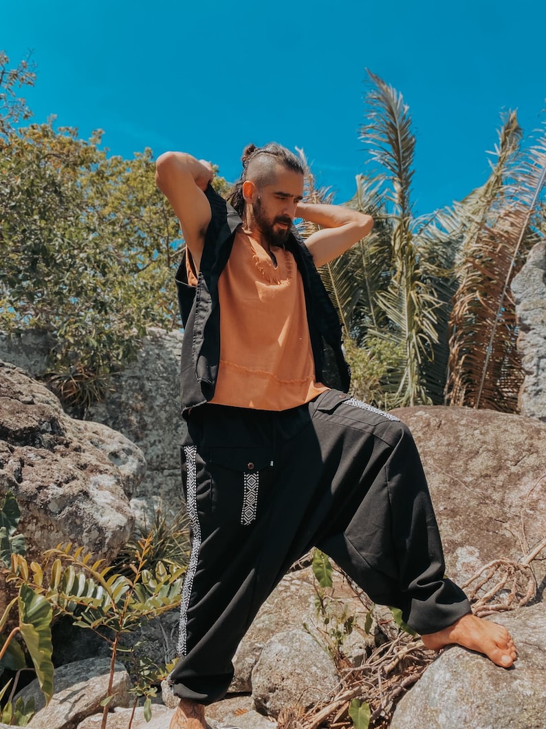 May include: A person wearing an orange top, black vest, and black pants with white patterned accents. The person is standing on rocks outdoors with a blue sky and greenery in the background.