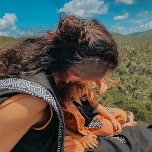 May include: A person wearing a black vest with white geometric patterns, and a child wearing an orange outfit with black accents. The background features a blue sky with clouds and a green landscape.