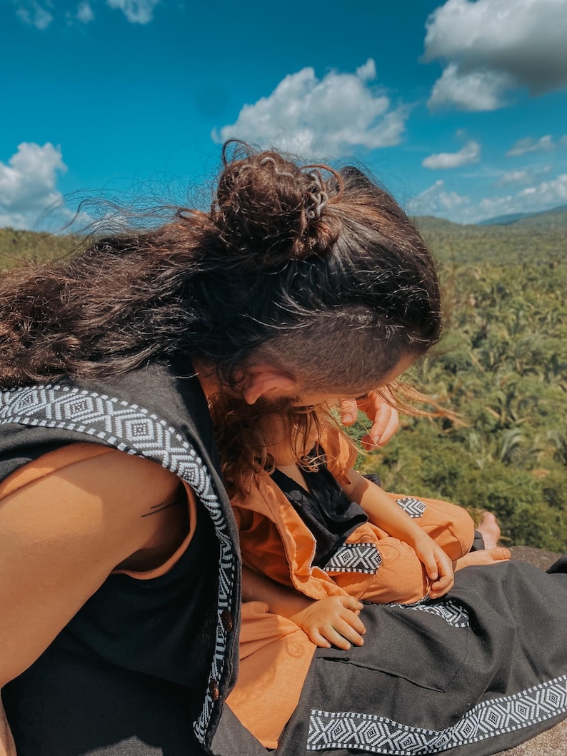 May include: A person wearing a black vest with white geometric patterns, and a child wearing an orange outfit with black accents. The background features a blue sky with clouds and a green landscape.