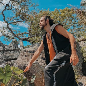 May include: A person wearing a black vest with white geometric trim over an orange shirt, paired with black pants featuring matching trim. The outfit is set against a backdrop of rocks, trees, and a bright blue sky.