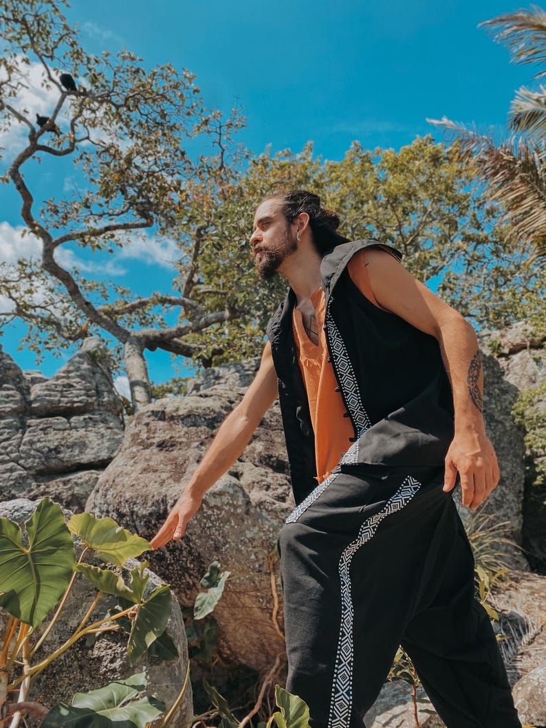 May include: A person wearing a black vest with white geometric trim over an orange shirt, paired with black pants featuring matching trim. The outfit is set against a backdrop of rocks, trees, and a bright blue sky.
