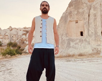 May include: A person wearing a cream-colored vest with brown buttons, a light blue shirt, and black harem pants. The person is standing on a dirt road with a rock formation in the background. The person is wearing flip-flops.