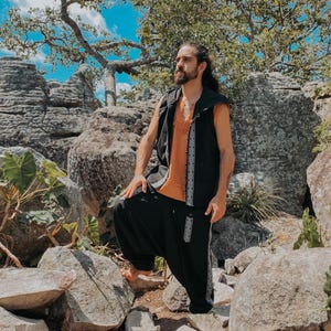 May include: A person wearing a black vest with white geometric trim over an orange shirt, paired with black harem pants featuring matching trim. The outfit is set against a backdrop of rocks and foliage under a bright blue sky.