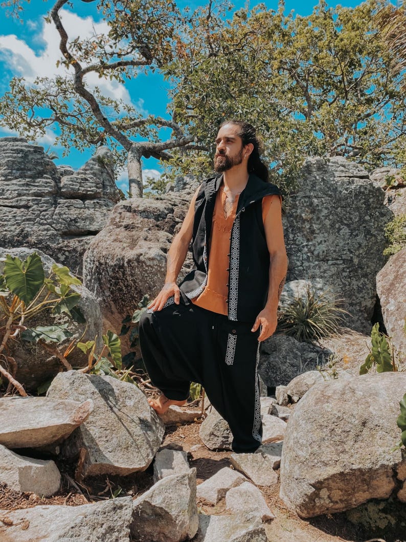 May include: A person wearing a black vest with white geometric trim over an orange shirt, paired with black harem pants featuring matching trim. The outfit is set against a backdrop of rocks and foliage under a bright blue sky.