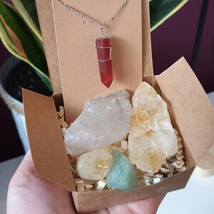 Könnte beinhalten: Geschenkbox mit einem roten Kristallanhänger und mehreren rohen Kristallen. Die Halskette hat einen roten Kristall, der in Silberdraht gewickelt ist. Die Box enthält auch klare, gelbe und blau-grüne Kristalle.