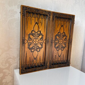Op de afbeelding: Een houten backgammonbord met ingewikkelde houtsnijwerk. Het bord is gemaakt van donker hout met een lichte houtnerf. De houtsnijwerk is donker van kleur en zeer gedetailleerd. Het bord is gesloten en klaar om te spelen.