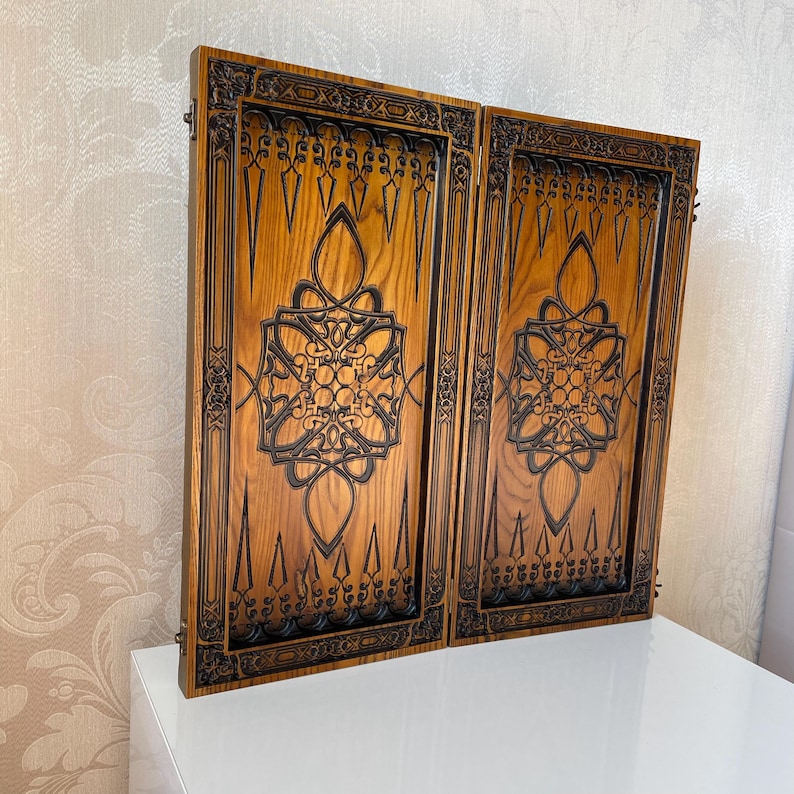 Op de afbeelding: Een houten backgammonbord met ingewikkelde houtsnijwerk. Het bord is gemaakt van donker hout met een lichte houtnerf. De houtsnijwerk is donker van kleur en zeer gedetailleerd. Het bord is gesloten en klaar om te spelen.