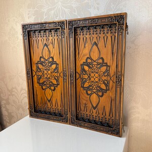 Op de afbeelding: Een houten backgammonbord met ingewikkelde houtsnijwerk. Het bord is gemaakt van lichtbruin hout met donkerbruine houtsnijwerk. Het bord is gesloten en de twee helften liggen tegenover elkaar.