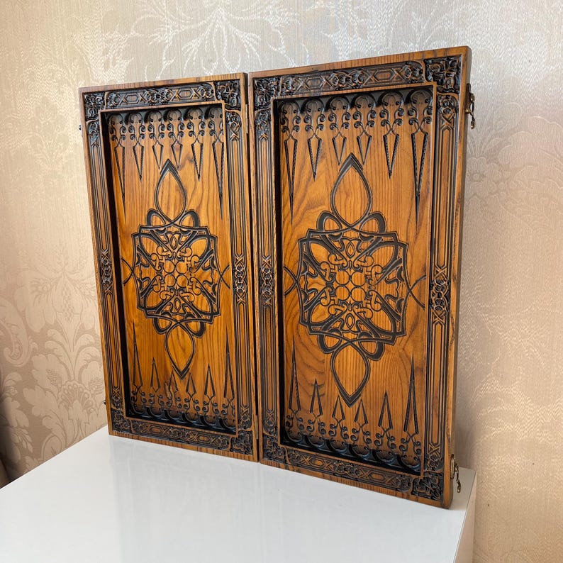 Op de afbeelding: Een houten backgammonbord met ingewikkelde houtsnijwerk. Het bord is gemaakt van lichtbruin hout met donkerbruine houtsnijwerk. Het bord is gesloten en de twee helften liggen tegenover elkaar.