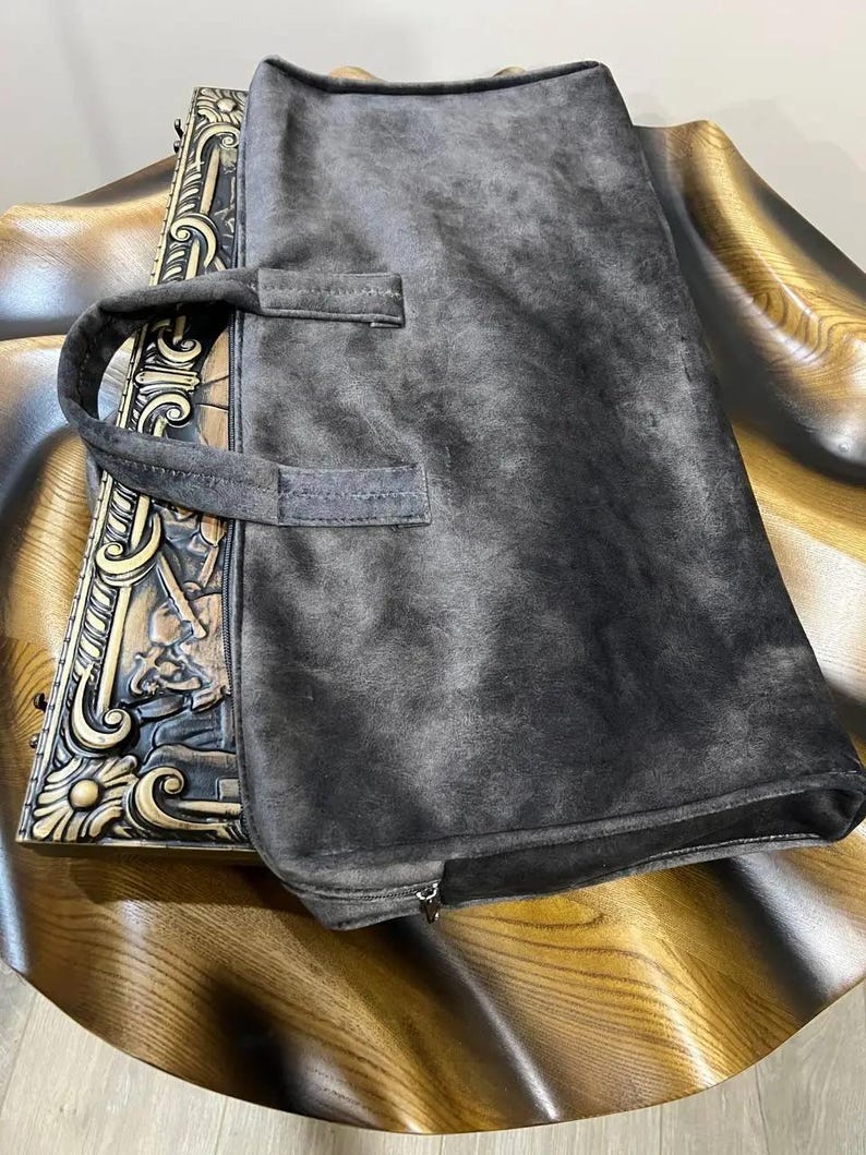 Op de afbeelding: Een grijze su&egrave;de draagtas met een ritssluiting en twee bovenste handvatten. De tas ligt op een houten oppervlak.