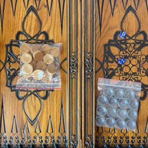 Op de afbeelding: Een houten backgammonbord met ingewikkelde houtsnijwerk. Het bord is in twee helften verdeeld, elk met een ander kleurenschema. De ene helft is bruin met zwarte accenten, de andere helft is zwart met bruine accenten. Het bord wordt geleverd met twee sets spelstukken, de ene set is bruin en de andere is zwart.