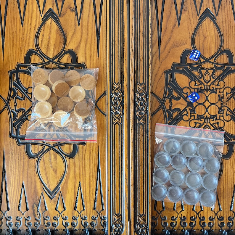 Op de afbeelding: Een houten backgammonbord met ingewikkelde houtsnijwerk. Het bord is in twee helften verdeeld, elk met een ander kleurenschema. De ene helft is bruin met zwarte accenten, de andere helft is zwart met bruine accenten. Het bord wordt geleverd met twee sets spelstukken, de ene set is bruin en de andere is zwart.