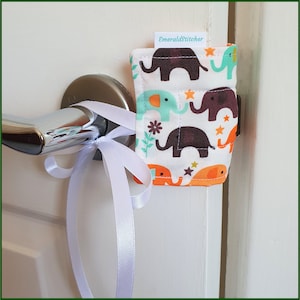 May include: A white door with a silver door handle. A white ribbon is tied around the handle. A small fabric doorstop with a white background and a pattern of colourful elephants and stars is attached to the door. The doorstop has a label that says "EmeraldStitcher".